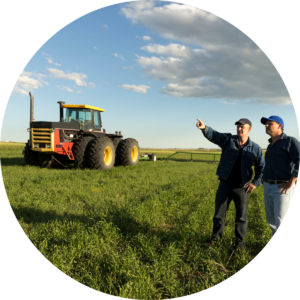 Two men stand in a field beside a tractor, surrounded by greenery and open space, engaged in conversation. One points to the horizon.